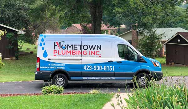 A Hometown Plumbing service van parked in a home's driveway. Green lawn on either side, and large trees in background.