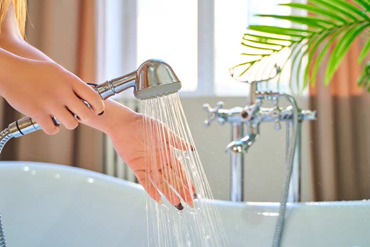 Person's hands holding a silver showerhead with water running, while other hand is testing the water. Soaking tub and sunny window in background.